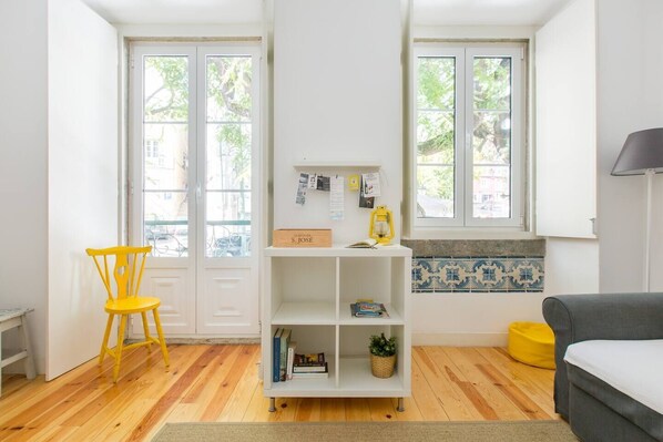 A room with a wooden floor, a yellow chair, a white bookshelf, a grey sofa, and a window with a view of trees.