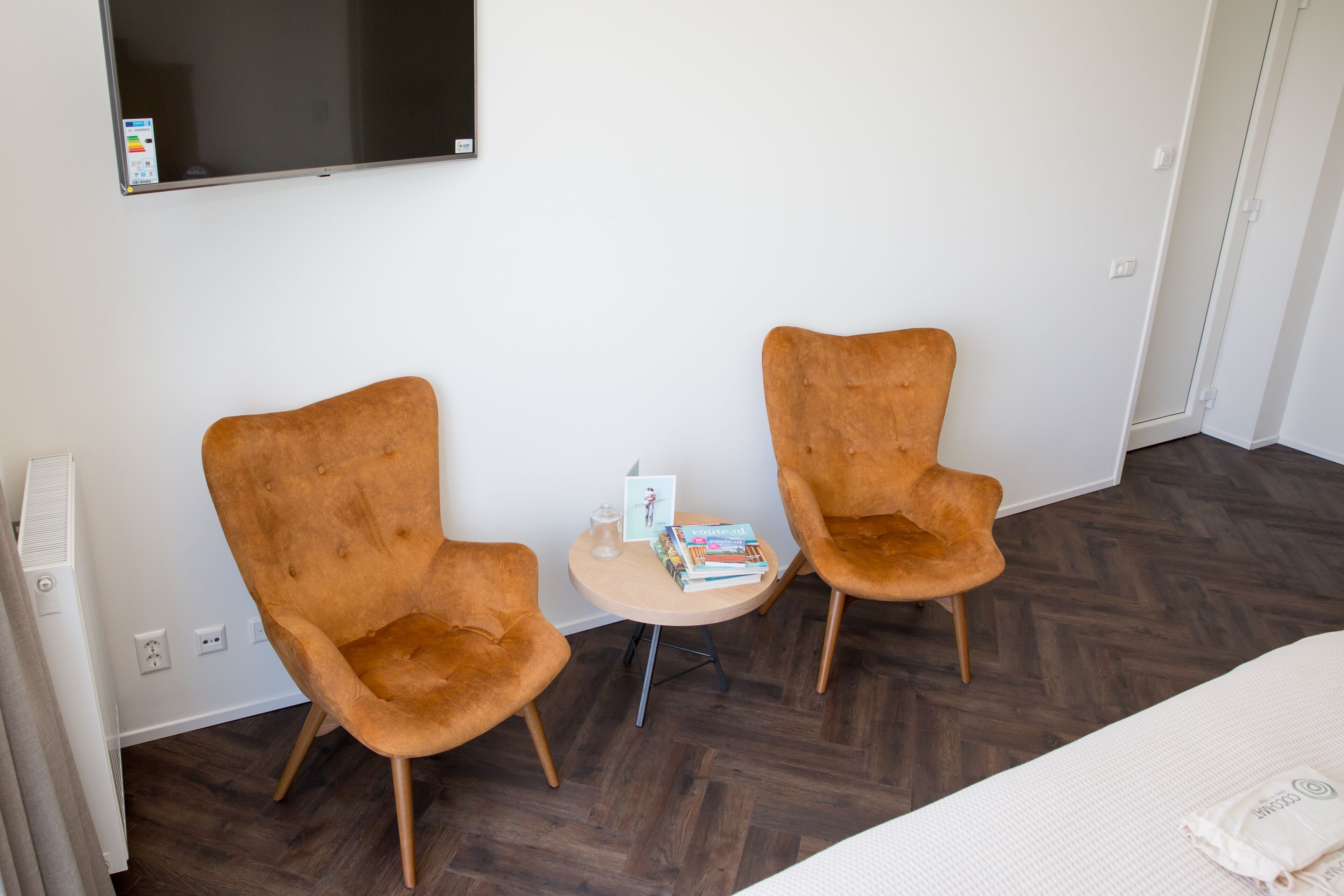 Une chambre d’hôtel avec deux fauteuils marron à revêtement en tissu, une petite table ronde sur laquelle sont posés des livres et une carte, un lit recouvert d’une couette blanche et un téléviseur à écran plat fixé au mur.