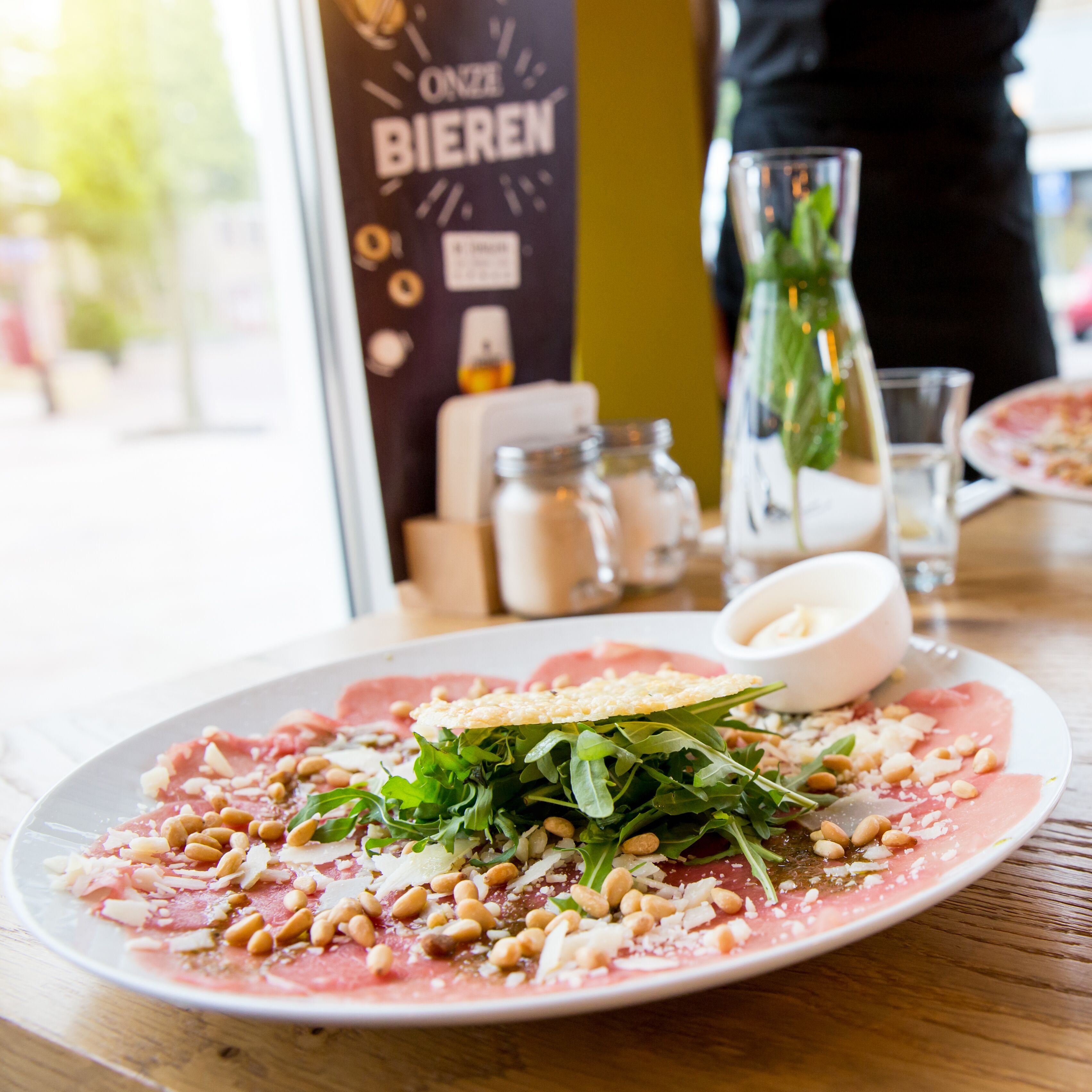 A plate of food with greens, nuts, and a slice of meat, a glass of water, and a bottle of olive oil on a table.