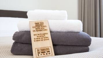 A stack of folded towels with a wooden tag that reads "TAKE THIS ORGANIC COTTON TOWEL ON ADVENTURE!" and a message about reusing towels to save water and the planet.