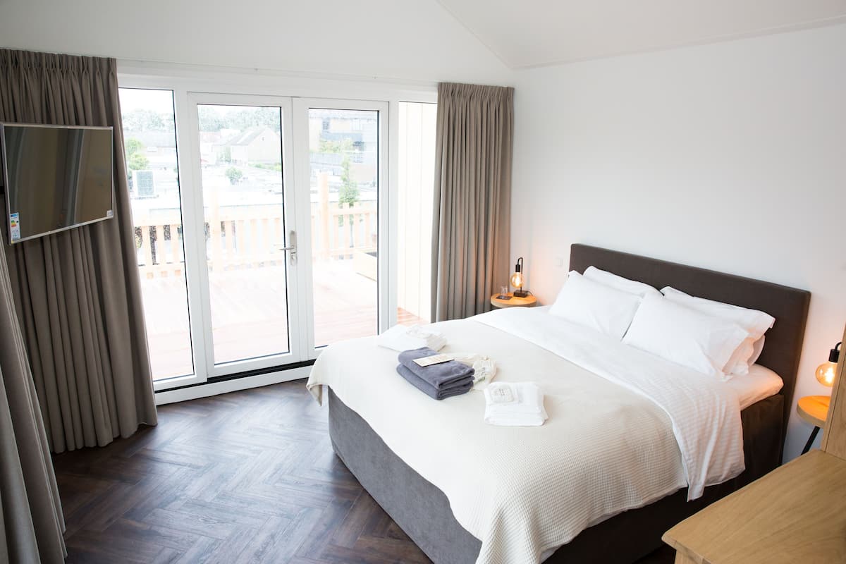 A hotel room with a bed, a TV mounted on the wall, a sliding glass door to the outside, and a bedside table with a lamp.
