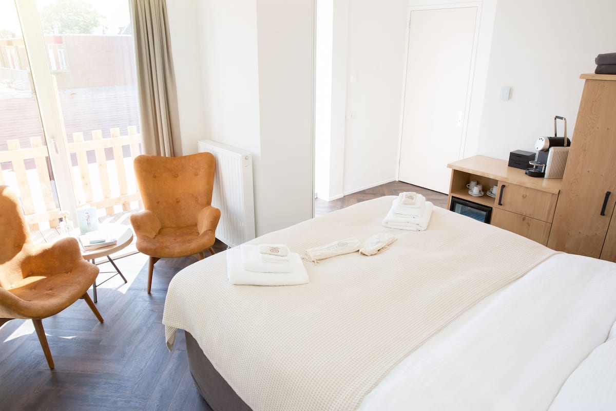 A hotel room with a large bed, two armchairs, a small table, a wooden cabinet, and a view of the outside.