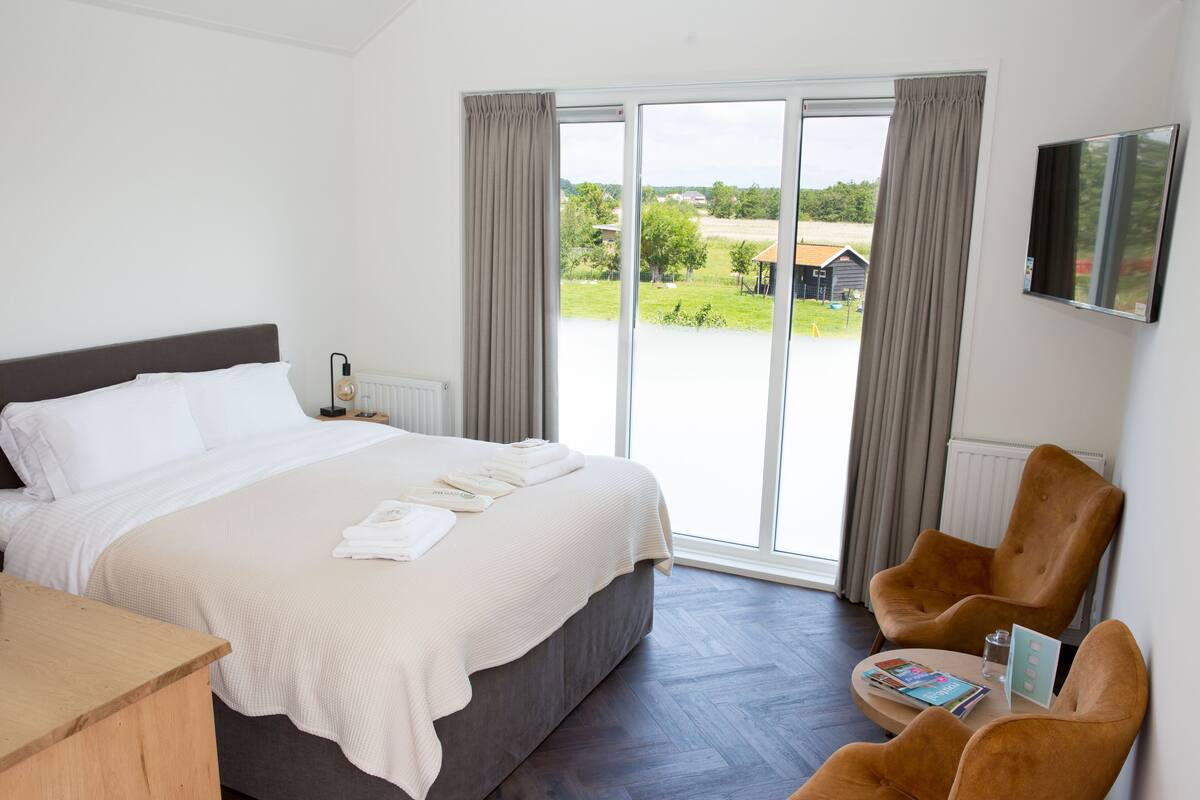 A hotel room with a large bed, a wooden desk, two armchairs, a TV, and a view of the outdoors.