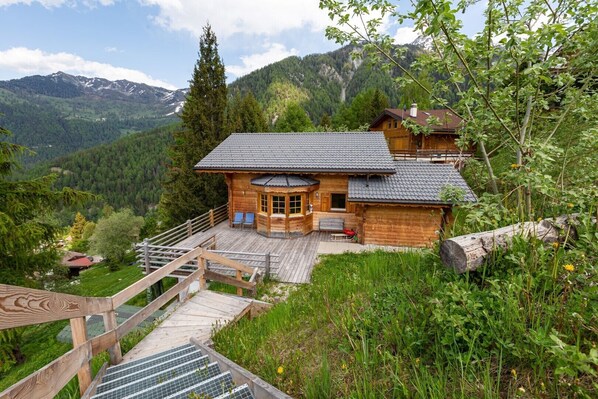 Un chalet de madera con balcón, rodeado de exuberante vegetación y montañas al fondo.