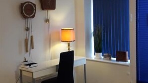 Una habitación con escritorio, silla y un reposapiés rojo. Hay una lámpara sobre el escritorio y una ventana con persianas.