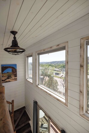 A room with white wooden walls, a ceiling fan, a large window with a view of a landscape, and a staircase.