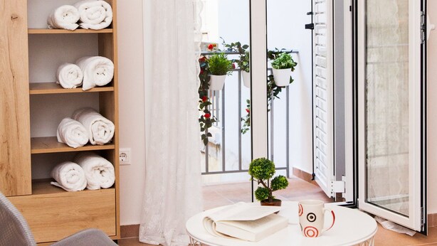 A room with a wooden shelf holding rolled towels, a round white table with a mug, a grey chair, and a view of a balcony with plants. - Cozy Studio Athens Downtown - Sleeps 3 (Athens)