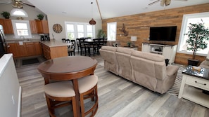 A modern kitchen with wooden cabinets, a dining area with a round table and chairs, a beige sofa, and a television. - Oceanside, Private Pool, Hot Tub, 410 ft to Beach Access! CP8, Moore Fun, Less Work (Corolla)