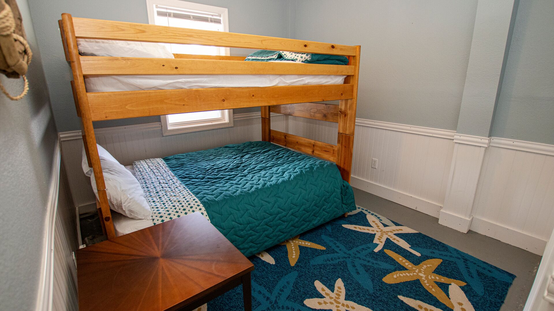 Quarto com beliche, cabeceira de madeira, uma cama individual com colcha azul-turquesa, uma pequena mesa de madeira e um tapete com padrão de estrelas-do-mar.