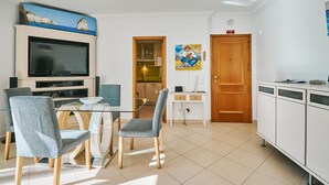 A modern living room with a TV, dining table, and chairs. There is a kitchen area with cabinets and a microwave. A painting hangs on the wall, and a door with an emergency sign is visible. - Apartment Meerlust with fantastic sea view and large balcony and garage (Armacao de Pera)