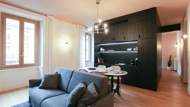 A modern living room with a grey sofa, wooden flooring, and a kitchen area with dark cabinets and a central island. - Cà Bèla - Vitruvio (Milan)