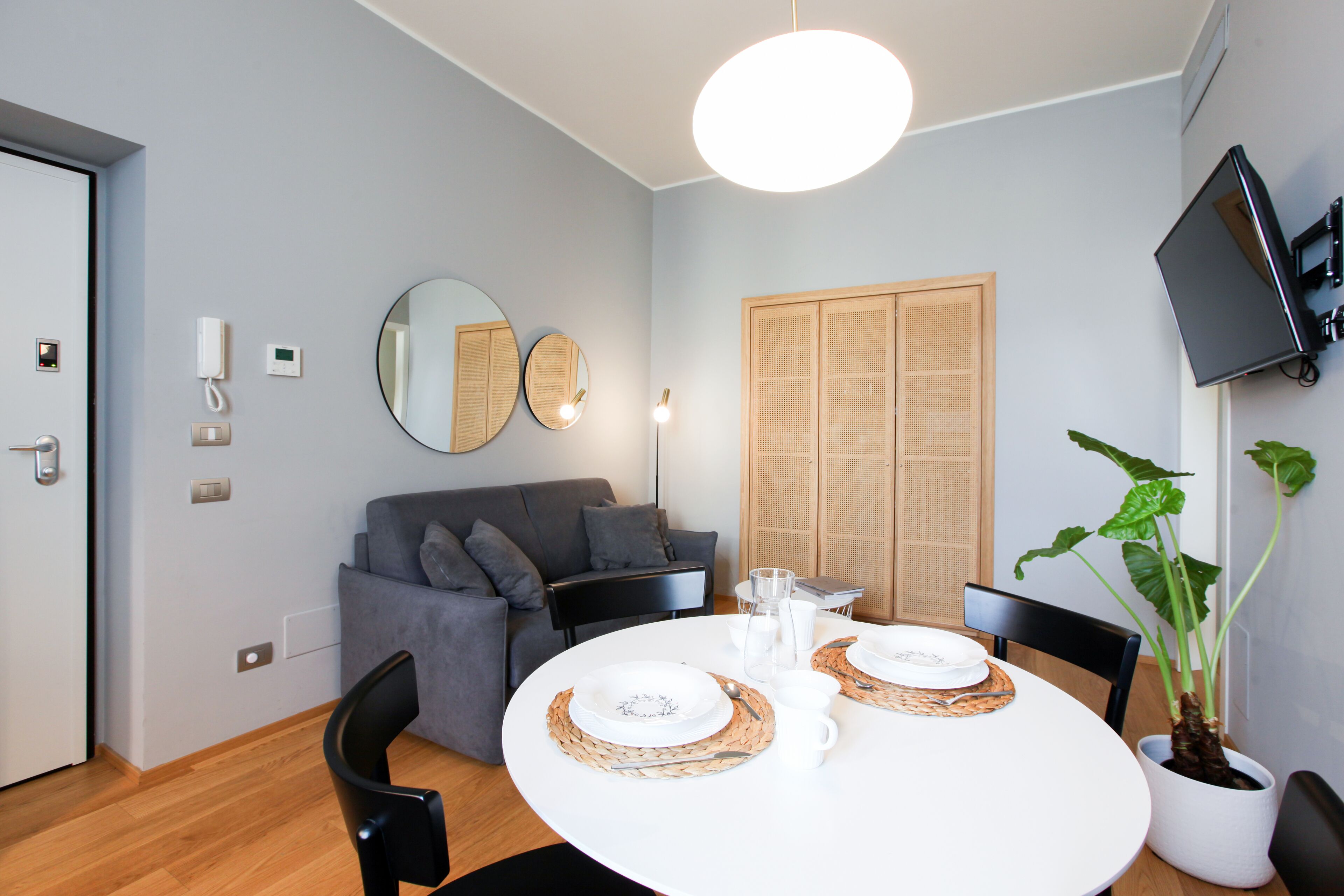 Un salon moderne avec une table à manger ronde préparée pour deux personnes, un canapé gris, un téléviseur fixé au mur et un grand miroir.