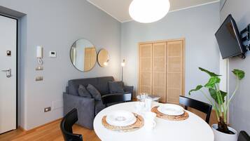 A modern living room with a round dining table set for two, a grey sofa, a television mounted on the wall, and a large mirror.