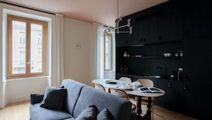 A modern living room with a grey sofa, a dining table with chairs, and a dark kitchen area. - Cà Bèla - Vitruvio (Milan)