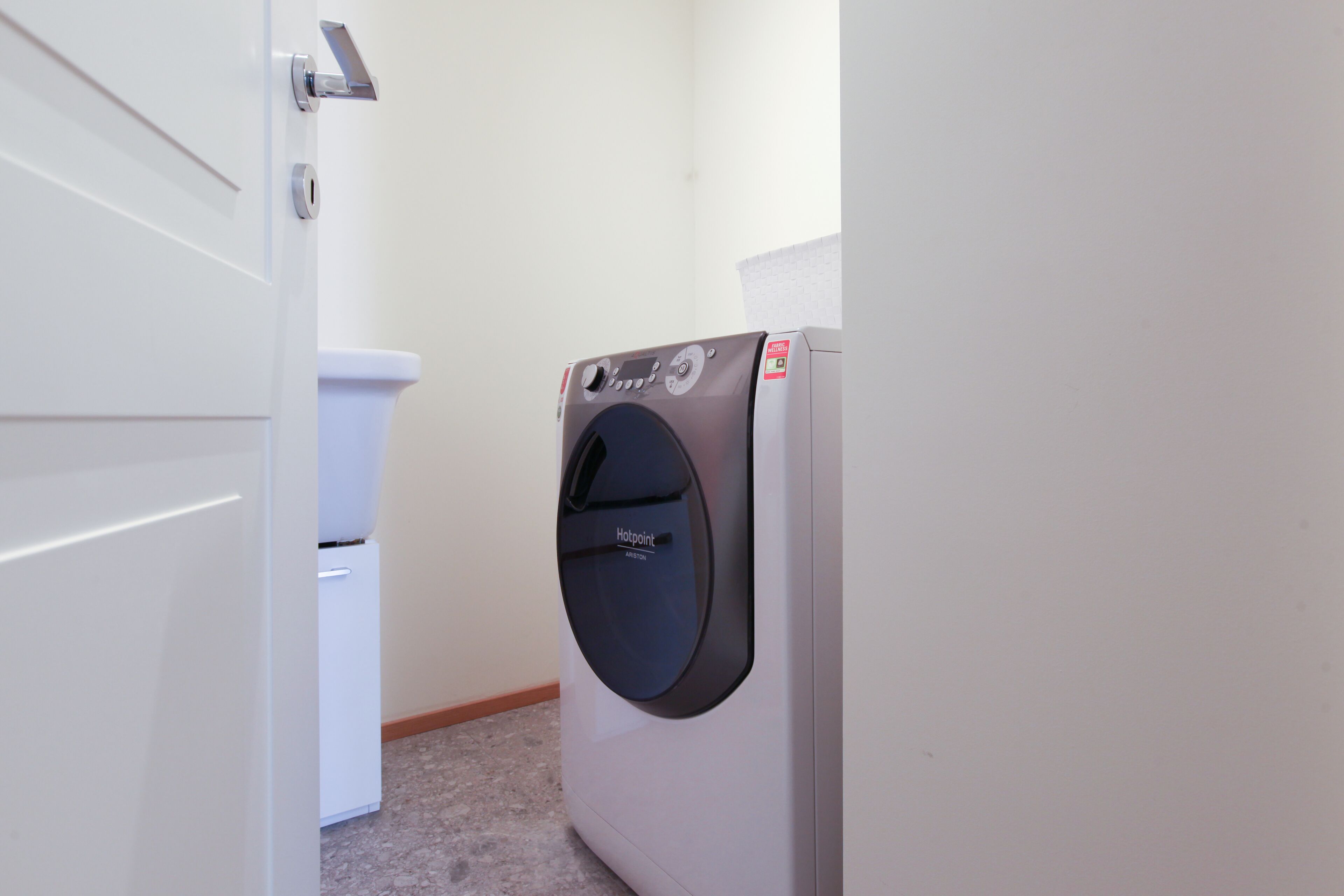 A compact, modern laundry room with a front-loading washer and dryer, a white toilet, and a door leading to another room.