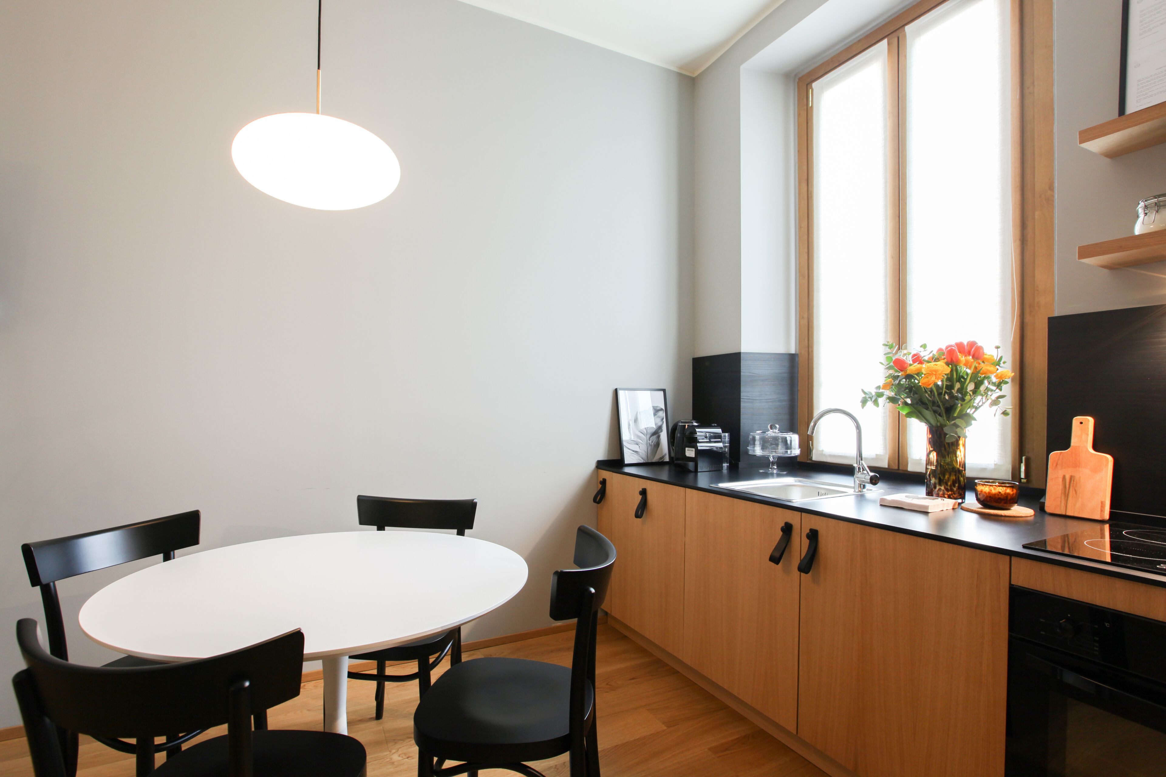 A modern kitchen with a white dining table, black chairs, wooden cabinets, and a window with natural light.