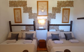 A hotel room with two beds, each with a headboard, pillows, and a small wooden stool. There is a wall-mounted light fixture between the beds, and artwork is displayed above the beds.