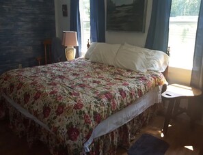 A bedroom with a floral bedspread, a wooden headboard, a nightstand, a lamp, and a window with curtains.