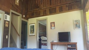 Une pièce avec un plafond en bois, un ventilateur de plafond, un lit recouvert d’une couverture bleue, un téléviseur posé sur un meuble en bois et un escalier menant à une autre pièce.