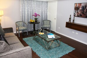Una sala de estar con una mesa de centro de vidrio, dos sillones estampados, una pequeña mesa redonda con un florero y fruta, y un televisor montado en la pared.