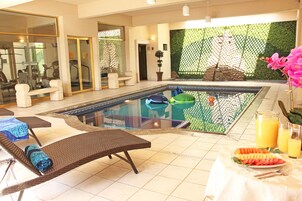 Un área de piscina con sillones de descanso, una mesa con frutas y bebidas, y equipo de ejercicios al fondo.