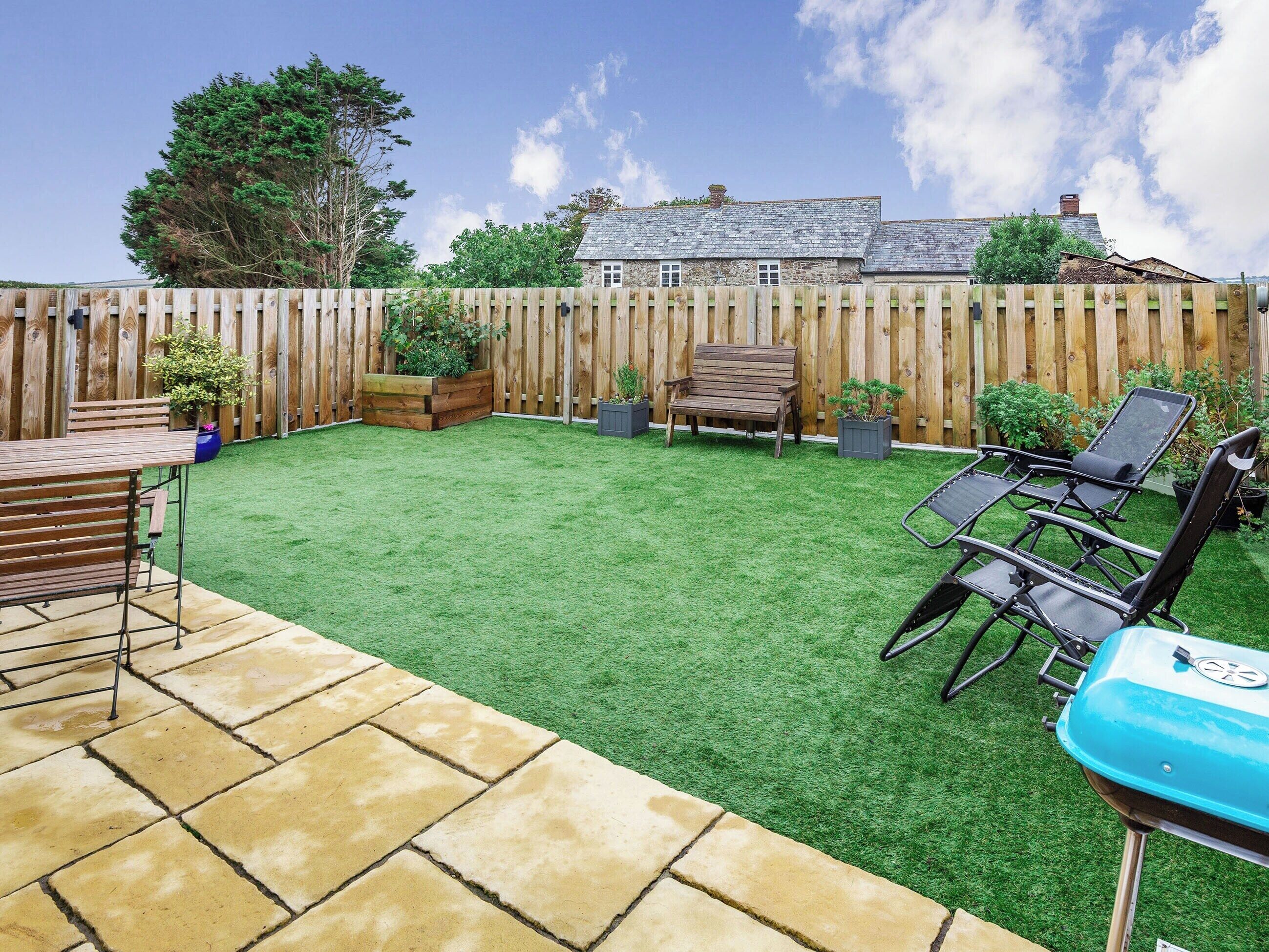 Un jardin avec une pelouse artificielle, une clôture en bois, un banc en bois, un barbecue et une table de patio.