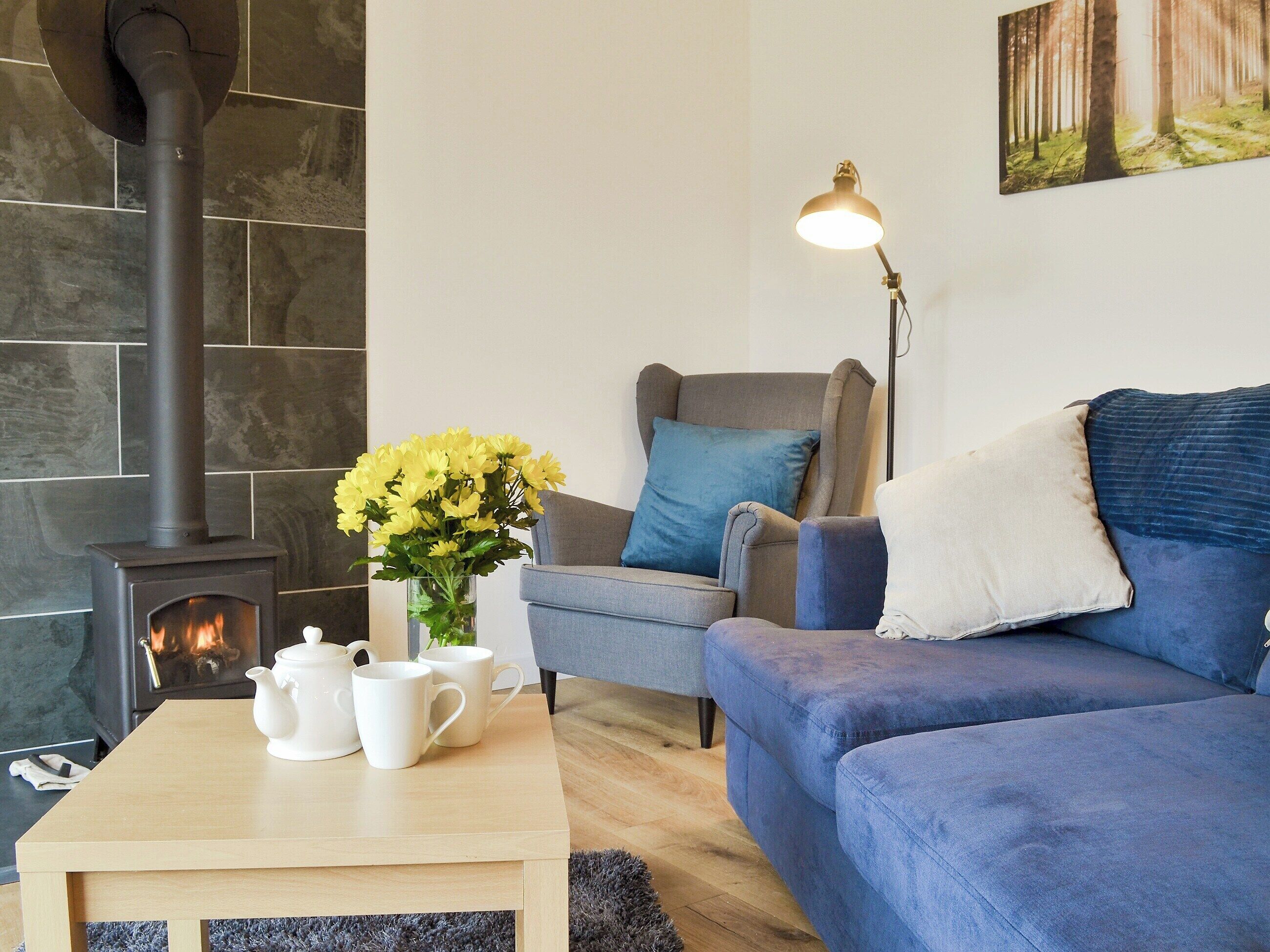 Una sala de estar acogedora con chimenea, un sofá azul, un sillón gris y una mesa con tazas blancas y un jarrón de flores amarillas.