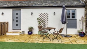 Uma casa com um alpendre ladrilhado, porta cinzenta, janela e um pequeno jardim com mesa e cadeiras.