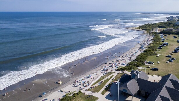 Une vue aérienne d’une plage avec une longue étendue de sable, un parking et des bâtiments aux toits distinctifs.