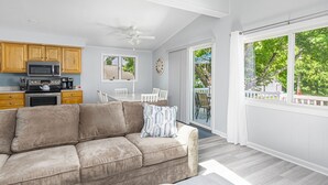 Un salon moderne avec un canapé beige, un coin repas avec des chaises blanches et une cuisine dotée de meubles et d’appareils électroménagers en bois.