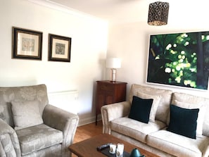 A living room with a sofa, armchair, and a coffee table. There are framed pictures on the wall and a lamp on a side table.