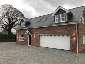 Une maison à deux étages en briques, avec une porte de garage blanche, un grand arbre en arrière-plan et une allée pavée.