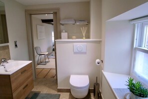 Un baño moderno con lavamanos, inodoro y zona de ducha blancos. Hay una planta pequeña a la derecha y una ventana al extremo derecho.