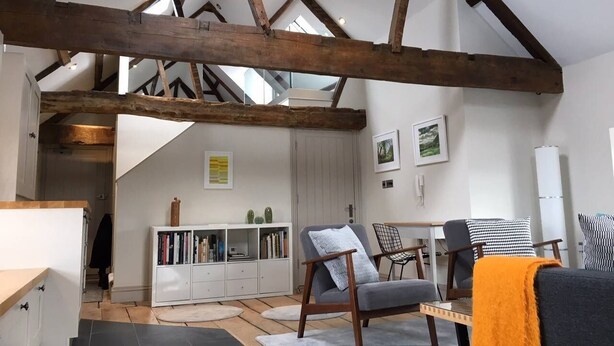 Un loft espacioso con vigas de madera a la vista, un rincón de descanso acogedor con sofá y sillones, una estantería blanca y una escalera que conduce a un nivel superior.