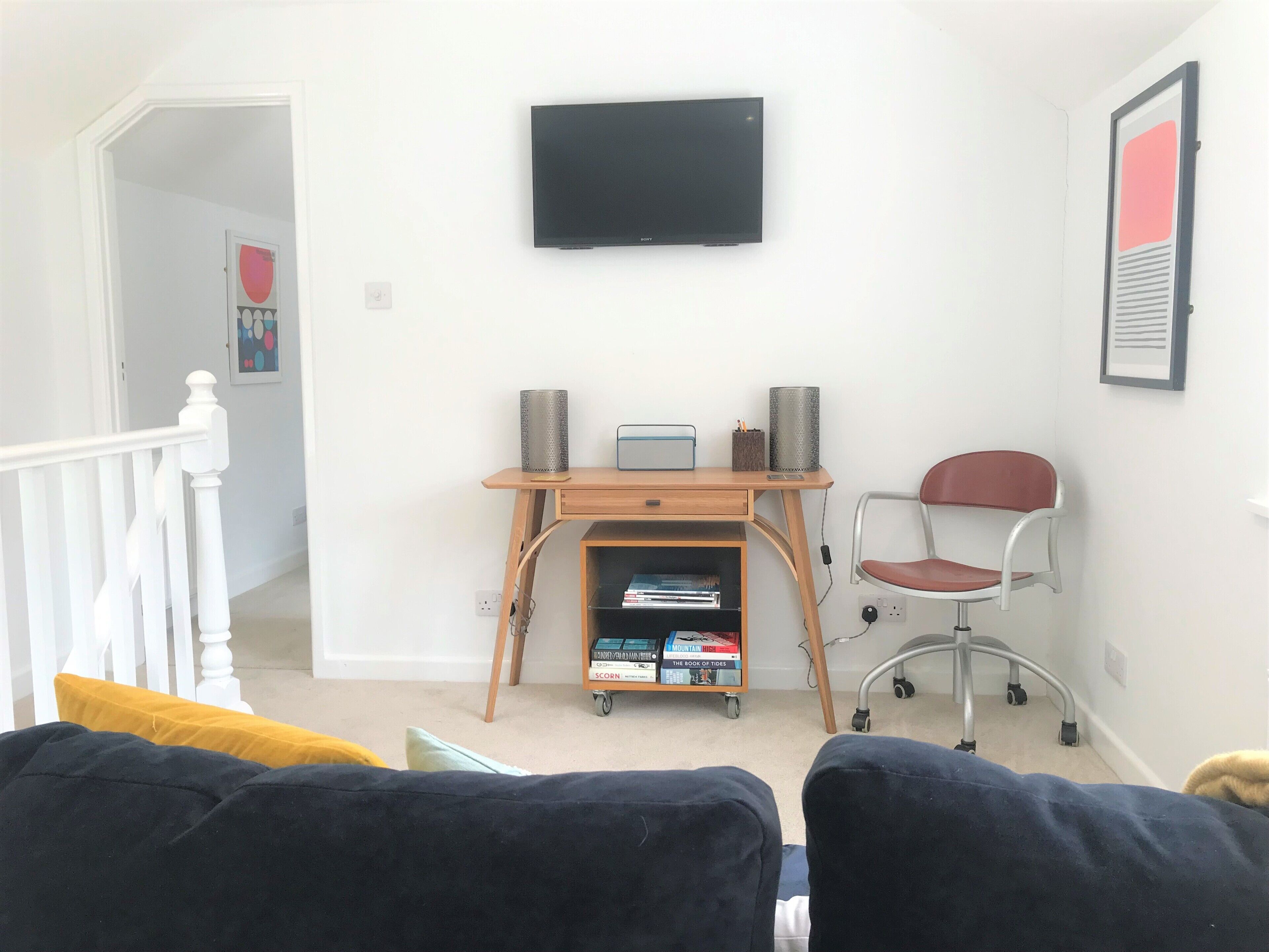 A living room with a TV mounted on the wall, a wooden desk with a chair, and a bookshelf.