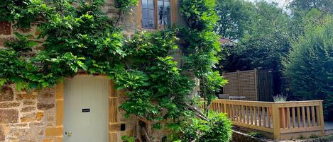 A stone house with a white door, a wooden deck, and lush greenery.