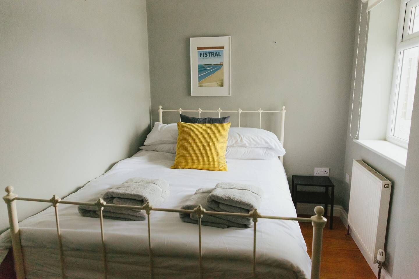 Un lit bien fait, avec des draps blancs et deux serviettes pliées, un oreiller jaune et un tableau encadré accroché au mur.
