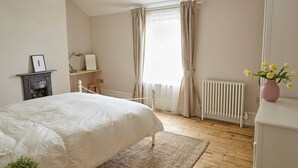 A bedroom with a bed, a radiator, a desk, a chair, a shelf with a plant, and a window with curtains.