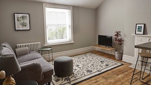 A living room with a grey sofa, a small round ottoman, a TV on a wooden stand, a fireplace, and a large window with blinds.