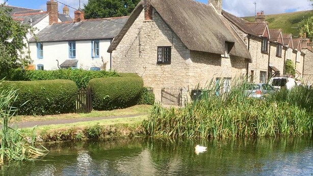 Une maison traditionnelle en pierre, avec un toit de chaume, située au bord d’un plan d’eau paisible, vraisemblablement un canal, agrémentée d’une haie bien entretenue et d’une clôture au premier plan.