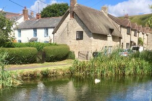 A traditional stone house with a thatched roof, situated beside a calm water body, likely a canal, with a well-maintained hedge and a fence in the foreground. - Idyllic Riverside Cottage in Dorset (Weymouth)