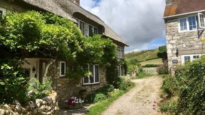 Une charmante maison en pierre avec un toit de chaume, entourée d’une végétation luxuriante et accessible par un chemin de gravier menant à un portail.