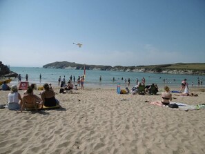 A beach with people sunbathing and swimming, a lifeguard tower, and a clear view of the sea and hills in the background.