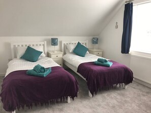 A twin bedroom with two beds, each with a purple bedspread and teal pillows. There are bedside tables with lamps and a window with curtains.