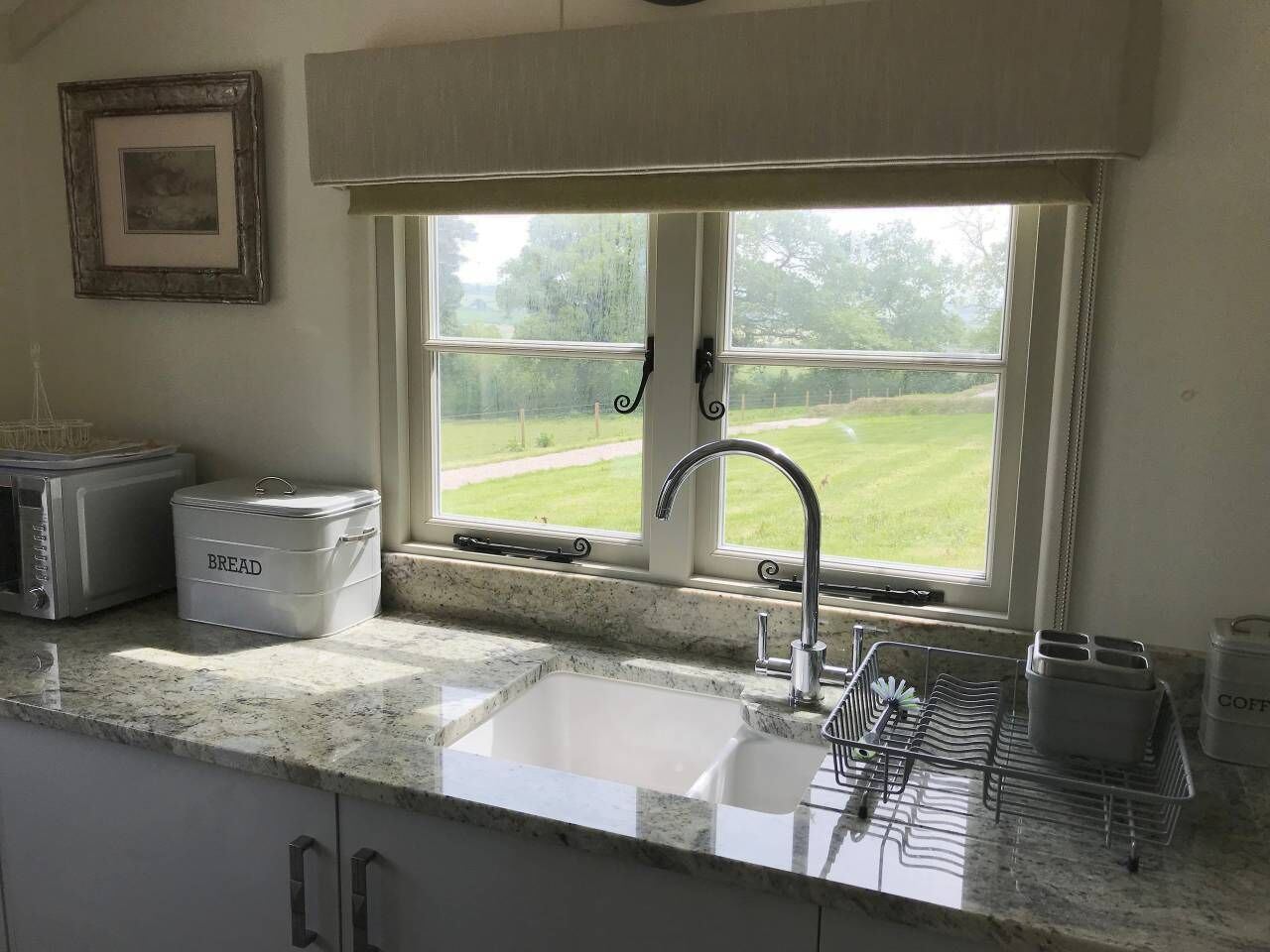 Una cocina con un lavamanos, un tostador, un recipiente para pan y una ventana con vistas a un paisaje verde.