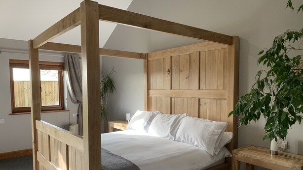 A four-poster bed with a wooden headboard, a bedside table with a lamp, and a plant on the right side. - Abbey View Cottage - Scandi Spa Hot Tub (Chard)