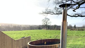 A wooden hot tub with a metal grill, set against a grassy field with a wooden fence and a tree in the background. - Abbey View Cottage - Scandi Spa Hot Tub (Chard)