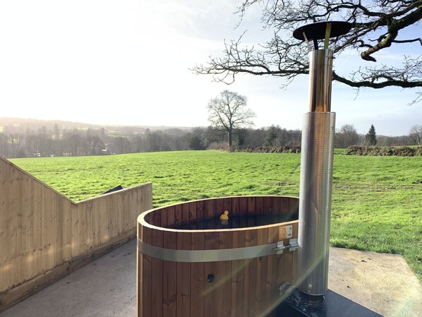 A wooden hot tub with a metal grill, set against a grassy field with a wooden fence and a tree in the background. - Abbey View Cottage - Scandi Spa Hot Tub (Chard)