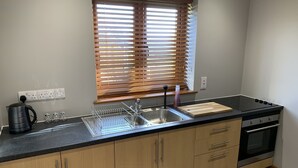 A modern kitchen with a stainless steel sink, wooden cabinets, a built-in oven, and a window with blinds. - Abbey View Cottage - Scandi Spa Hot Tub (Chard)