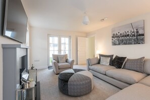 A modern living room with a sofa, armchair, ottoman, and a flat-screen TV mounted on the wall. - Stylish & Modern 3 Bedroom Home (Bangor)